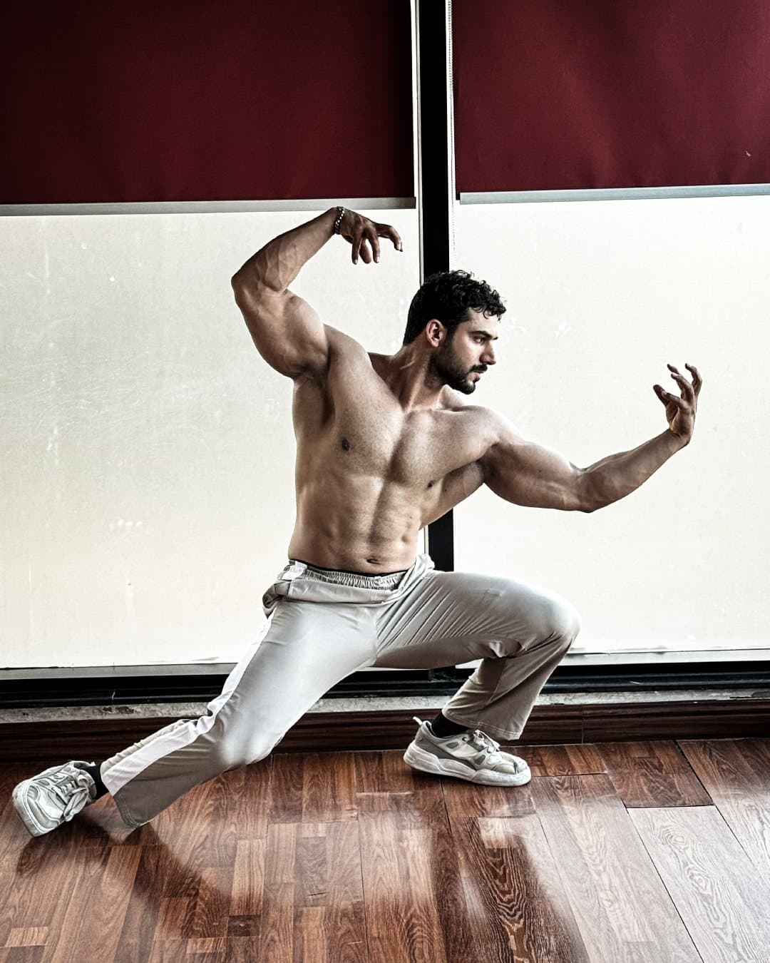 Expressive lunge pose, arms raised, in front of burgundy blinds
